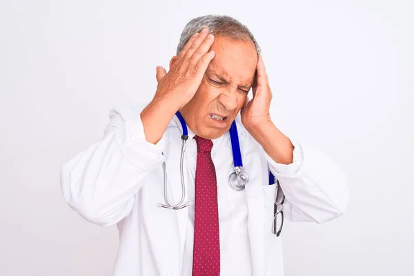 ストレスのため頭の痛みに頭を抱えて孤立した白い背景の上に立つ聴診器を身に着けている先輩の灰色の髪の医者の男性 苦しむ片頭痛 — ストック写真