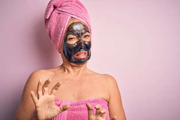 Middle age brunette woman wearing beauty black face mask over isolated pink background disgusted expression, displeased and fearful doing disgust face because aversion reaction.
