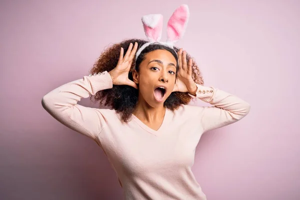 ピンクの背景にウサギの耳を身に着けているアフロの髪を持つ若いアフリカ系アメリカ人の女性明るい顔を示す手で竹を覗かせる笑顔 驚きと興奮した — ストック写真