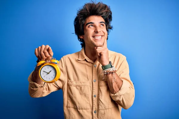 Jovem Homem Bonito Segurando Despertador Sobre Fundo Azul Isolado Rosto — Fotografia de Stock