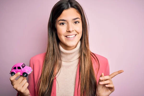 Young beautiful girl holding small car toy standing over isolated pink background very happy pointing with hand and finger to the side