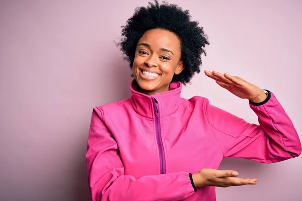 Young African American afro sportswoman with curly hair wearing sportswear doin sport gesturing with hands showing big and large size sign, measure symbol. Smiling looking at the camera. Measuring concept.
