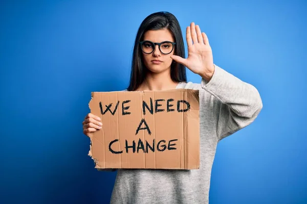 Young beautiful brunette activist woman holding banner protesting to change with open hand doing stop sign with serious and confident expression, defense gesture