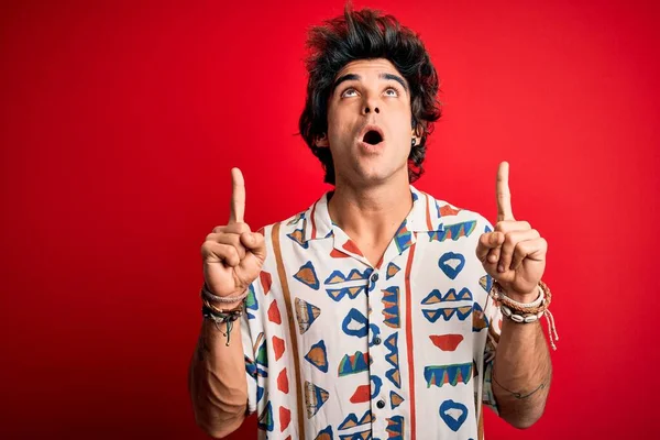 stock image Young handsome man on vacation wearing summer shirt over isolated red background amazed and surprised looking up and pointing with fingers and raised arms.