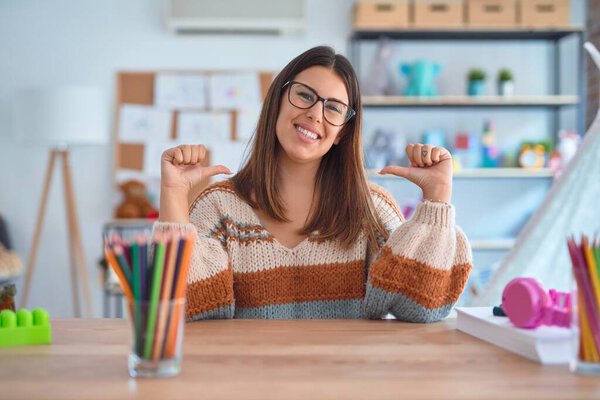Young beautiful teacher woman wearing sweater and glasses sitting on desk at kindergarten looking confident with smile on face, pointing oneself with fingers proud and happy.