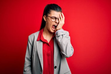 Young beautiful brunette businesswoman wearing jacket and glasses over red background Yawning tired covering half face, eye and mouth with hand. Face hurts in pain.