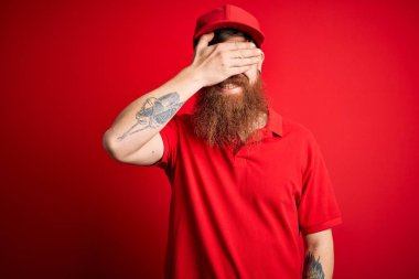 Gözlüklü ve kırmızı şapkalı genç teslimatçı izole edilmiş arka planda gülümsüyor ve yüzünde şaşkınlıktan gözleri kapayarak gülüyor. Kör kavram.