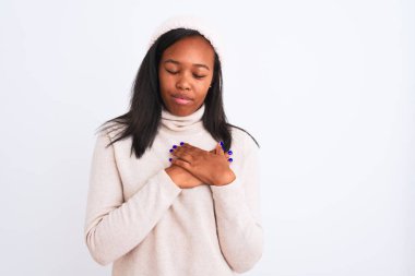 Beautiful young african american woman wearing turtleneck sweater and winter hat smiling with hands on chest with closed eyes and grateful gesture on face. Health concept.