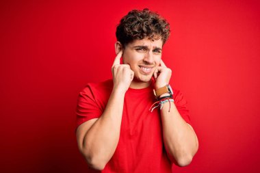 Sarışın, kıvırcık saçlı, kırmızı arka plan üzerine gündelik tişört giyen, yüksek sesli müzik sesi için rahatsız edici ifadeleri olan parmaklarıyla kulaklarını kapatan genç, yakışıklı bir adam. Sağır kavramı.