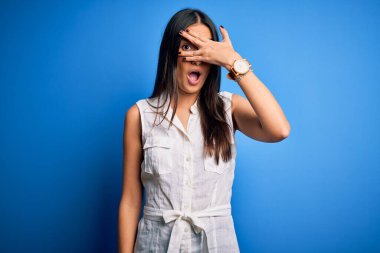 Young beautiful brunette woman wearing casual dress over isolated blue background peeking in shock covering face and eyes with hand, looking through fingers with embarrassed expression.