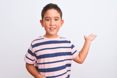 Beautiful kid boy wearing casual striped t-shirt standing over isolated white background smiling cheerful presenting and pointing with palm of hand looking at the camera.
