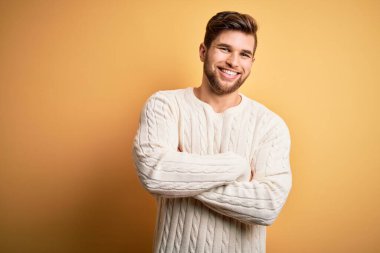 Sarışın, sakallı, mavi gözlü, sarı arka planda beyaz süveter giyen, kollarını kavuşturup kameraya bakan genç bir adam. Pozitif kişi.