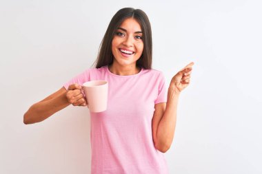 Genç ve güzel bir kadın, izole edilmiş beyaz arka planda duran pembe bir fincan kahve içerken mutlu bir şekilde elini ve parmağını kenarı işaret ediyor.