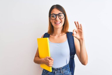 Beautiful redhead student woman wearing a backpack and book doing ok sign with fingers, excellent symbol