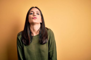 Young brunette woman with blue eyes wearing green casual sweater over yellow background looking at the camera blowing a kiss on air being lovely and sexy. Love expression.