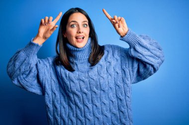 Young brunette woman with blue eyes wearing casual turtleneck sweater smiling amazed and surprised and pointing up with fingers and raised arms.