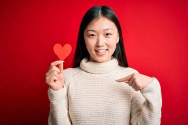 Young asian woman holding romantic red heart paper shape over red isolated background with surprise face pointing finger to himself