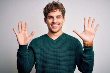 Sarışın, kıvırcık saçlı, beyaz arka planda yeşil süveter giyen, on numaralı parmaklarıyla onu gösteren, kendinden emin ve mutlu gülümseyen genç bir adam..