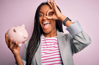 Young african american business woman saving money on piggy bank over isolated background with happy face smiling doing ok sign with hand on eye looking through fingers