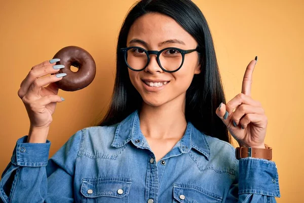 Young Beautiful Chinese Woman Holding Chocolate Donut Isolated Yellow Background — Stockfoto