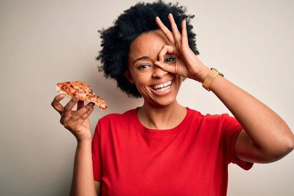Young African American afro woman with curly hair eating slice of delicious Italian pizza with happy face smiling doing ok sign with hand on eye looking through fingers