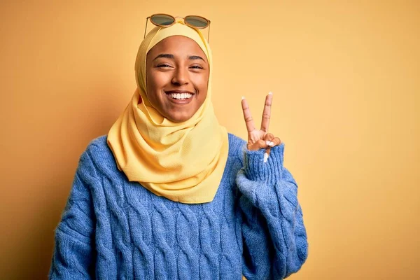Sarı başörtüsü ve güneş gözlüğü takan, kameraya gülümseyen, zafer işareti yapan Afro-Amerikan genç bayan öğrenci. İki numara..