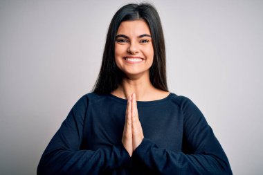 Young beautiful brunette woman wearing casual sweater standing over white background praying with hands together asking for forgiveness smiling confident.