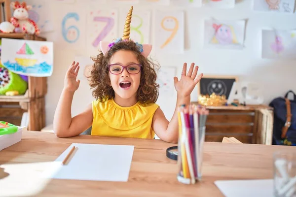 幼稚園の机の上に座っている眼鏡とユニコーンのダイエムを身に着けている美しい幼児は 狂気を祝い 腕を上げて開いた目が興奮して叫んで成功したことに驚いています 勝者のコンセプト — ストック写真