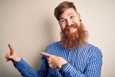 Yakışıklı, kızıl saçlı, sakallı bir iş adamı izole edilmiş arka planda duruyor gülümsüyor ve kameraya bakıyor. İki eliyle ve parmaklarıyla işaret ediyor..