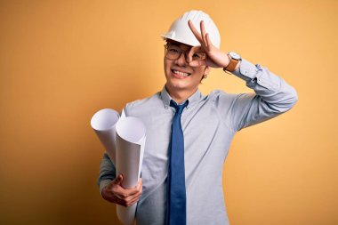 Young handsome chinese architect man wearing safety helmet holding blueprints with happy face smiling doing ok sign with hand on eye looking through fingers