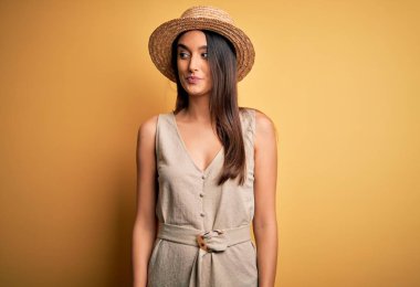Young beautiful brunette woman on vacation wearing casual dress and hat smiling looking to the side and staring away thinking.
