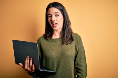 Young brunette woman with blue eyes working using computer laptop over yellow background winking looking at the camera with sexy expression, cheerful and happy face.