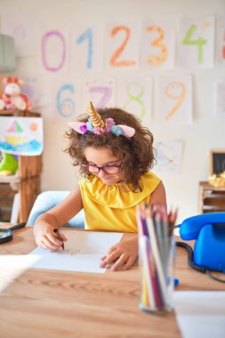 Gözlüklü ve tek boynuzlu at diademi takmış güzel bir çocuk. Anaokulunda kağıt ve keçeli kalemle çizim yapıyor.
