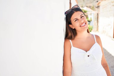 Güzel kız beyaz duvara yaslanıyor, genç arkadaş canlısı kadın mutlu bir yaz gününde gülümsüyor.