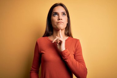 Young beautiful brunette woman wearing casual t-shirt standing over yellow background Thinking concentrated about doubt with finger on chin and looking up wondering