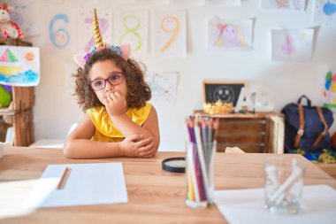 Beautiful toddler wearing glasses and unicorn diadem sitting on desk at kindergarten looking stressed and nervous with hands on mouth biting nails. Anxiety problem.