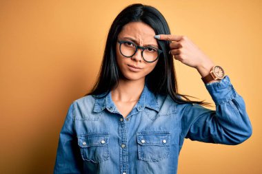 Young beautiful chinese woman wearing casual denim shirt over isolated yellow background pointing unhappy to pimple on forehead, ugly infection of blackhead. Acne and skin problem