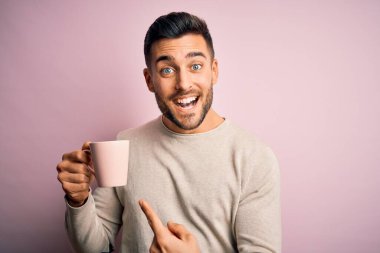 Young handsome man drinking a cup of hot coffee over pink isolated background very happy pointing with hand and finger