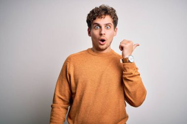 Young blond handsome man with curly hair wearing casual sweater over white background Surprised pointing with hand finger to the side, open mouth amazed expression.