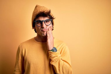 Young handsome man wearing casual t-shirt and glasses over isolated yellow background touching mouth with hand with painful expression because of toothache or dental illness on teeth. Dentist