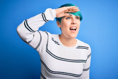 Young beautiful woman with blue fashion hair wearing casual sweater standing at studio very happy and smiling looking far away with hand over head. Searching concept.