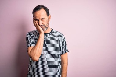 Sıradan çizgili tişört giyen orta yaşlı beyaz bir adam izole edilmiş pembe arka planda duruyor. Kollarını kavuşturup depresyondan sıkılmış ve yorgun görünüyor..