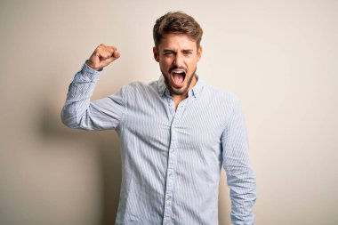 Sakallı, çizgili tişörtlü genç bir adam beyaz arka planda kızgın ve kızgın duruyor. Öfkeyle bağırırken yumruğunu kaldırıyor. Öfke ve agresif kavram.