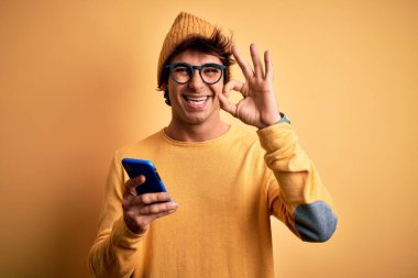 Young handsome man using smartphone wearing glasses over isolated yellow background doing ok sign with fingers, excellent symbol