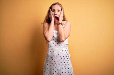 Young beautiful blonde woman on vacation wearing summer dress over yellow background afraid and shocked, surprise and amazed expression with hands on face