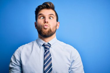 Young blond businessman with beard and blue eyes wearing elegant shirt and tie standing making fish face with lips, crazy and comical gesture. Funny expression.
