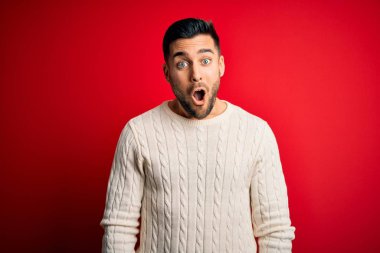 Genç yakışıklı adam, günlük beyaz süveter giyiyor. Kırmızı arka planda duruyor. Şaşırmış ve korkmuş bir ifade, korku ve heyecanlı bir yüz..