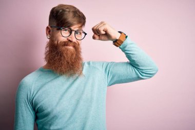 Pembe arka planda gözlüklü, sakallı yakışıklı kızıl saçlı güçlü kol kası olan, kendinden emin ve gururlu bir adam.