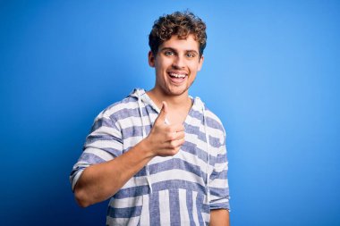 Young blond handsome man with curly hair wearing casual striped sweatshirt doing happy thumbs up gesture with hand. Approving expression looking at the camera showing success.