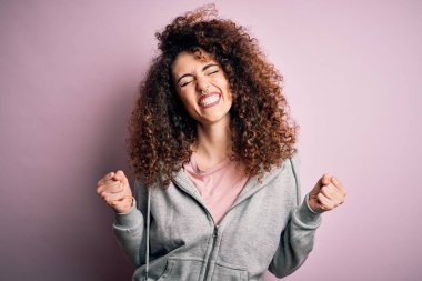 Genç, esmer, kıvırcık saçlı, piercing takan, spor giyinen, çok mutlu ve heyecanlı genç bir bayan. Kazanan jestler yapıyor, kollarını kaldırıp gülümsüyor ve başarı için bağırıyor. Kutlama konsepti.